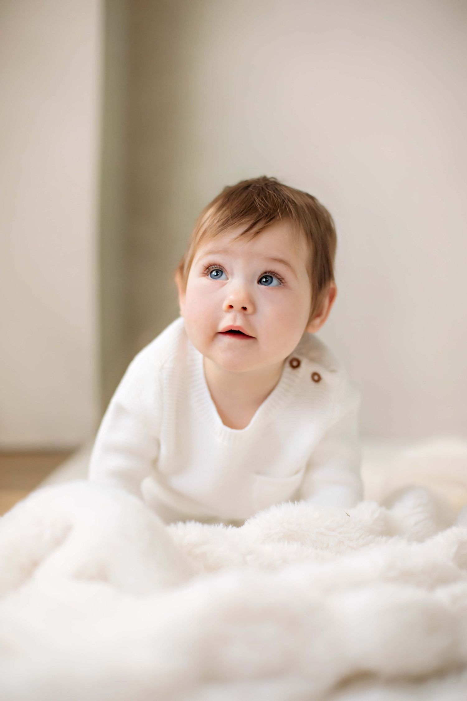 Baby in white sweater laying on soft white blanket with neutral background.