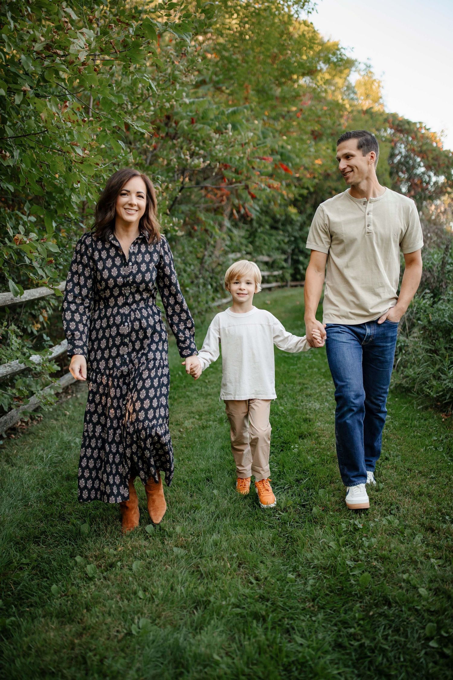Family of three walking hand in hand on grassy path during sunset.