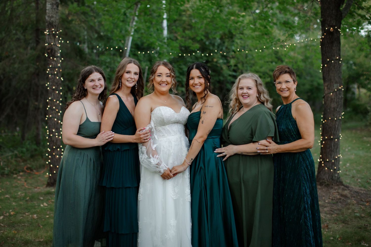 Wedding party wearing dark teal bridesmaid dresses poses outdoors in a wooded setting at sunset.