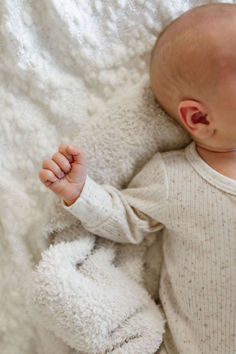 Close-up of tiny baby hand and arm in cream ribbed sweater against white textured blanket.