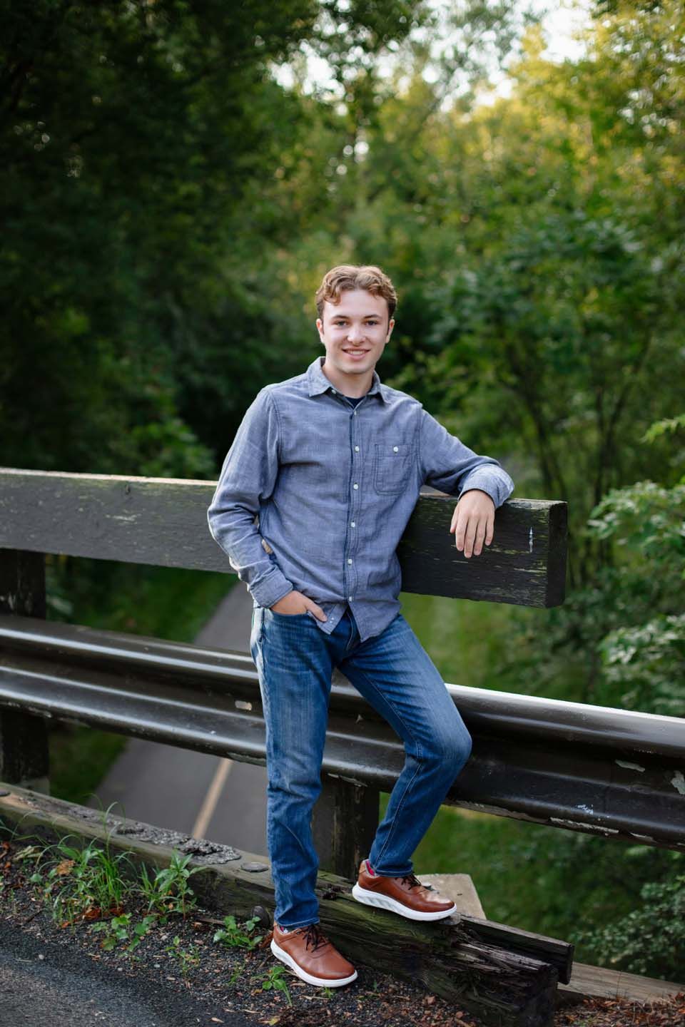 Casual portrait of someone posing on wooden railing in blue button-down shirt and jeans in outdoor park setting.