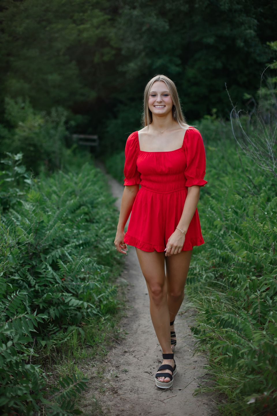 Person walking along a dirt trail wearing a red dress and sandals surrounded by lush greenery.