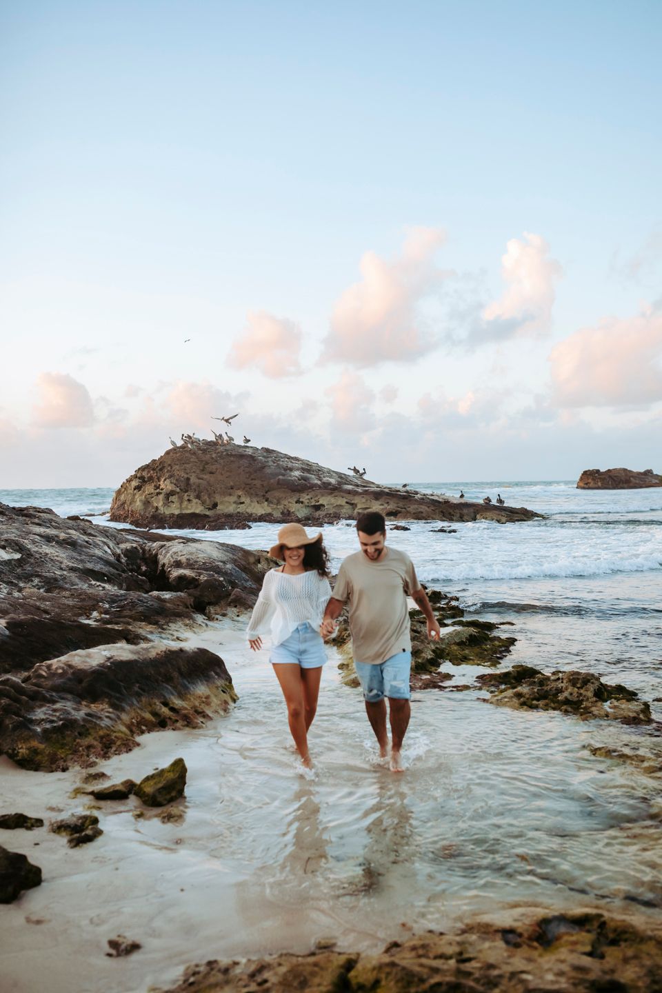 A couple walking hand in hand through shallow ocean water at sunset among rocky coastal formations in Tulum.