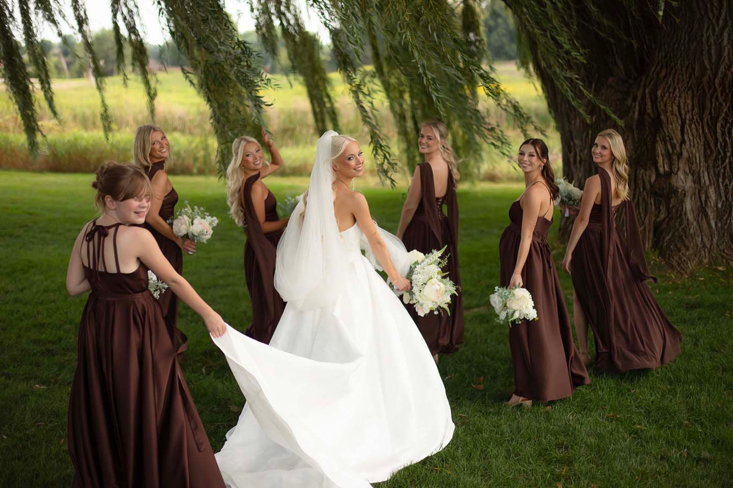 Bride in white gown with veil surrounded by bridesmaids in brown dresses holding white bouquets under a willow tree.