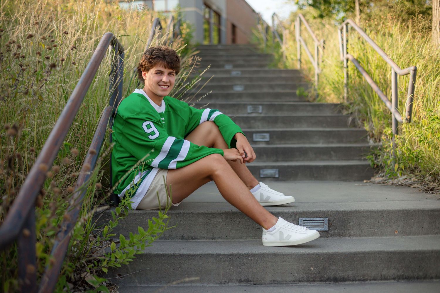 Teen boy in green jersey number 9 sitting on outdoor concrete steps, smiling in golden hour light.