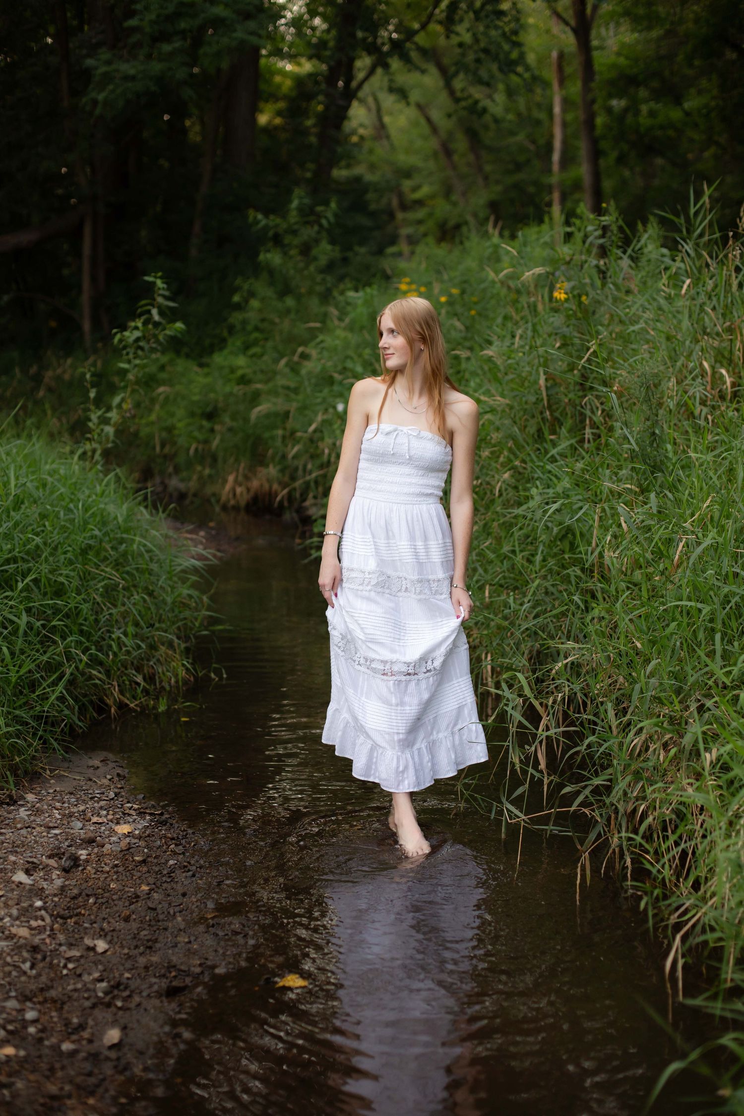 Young woman in white sundress wading barefoot in a shallow forest creek surrounded by lush green vegetation.