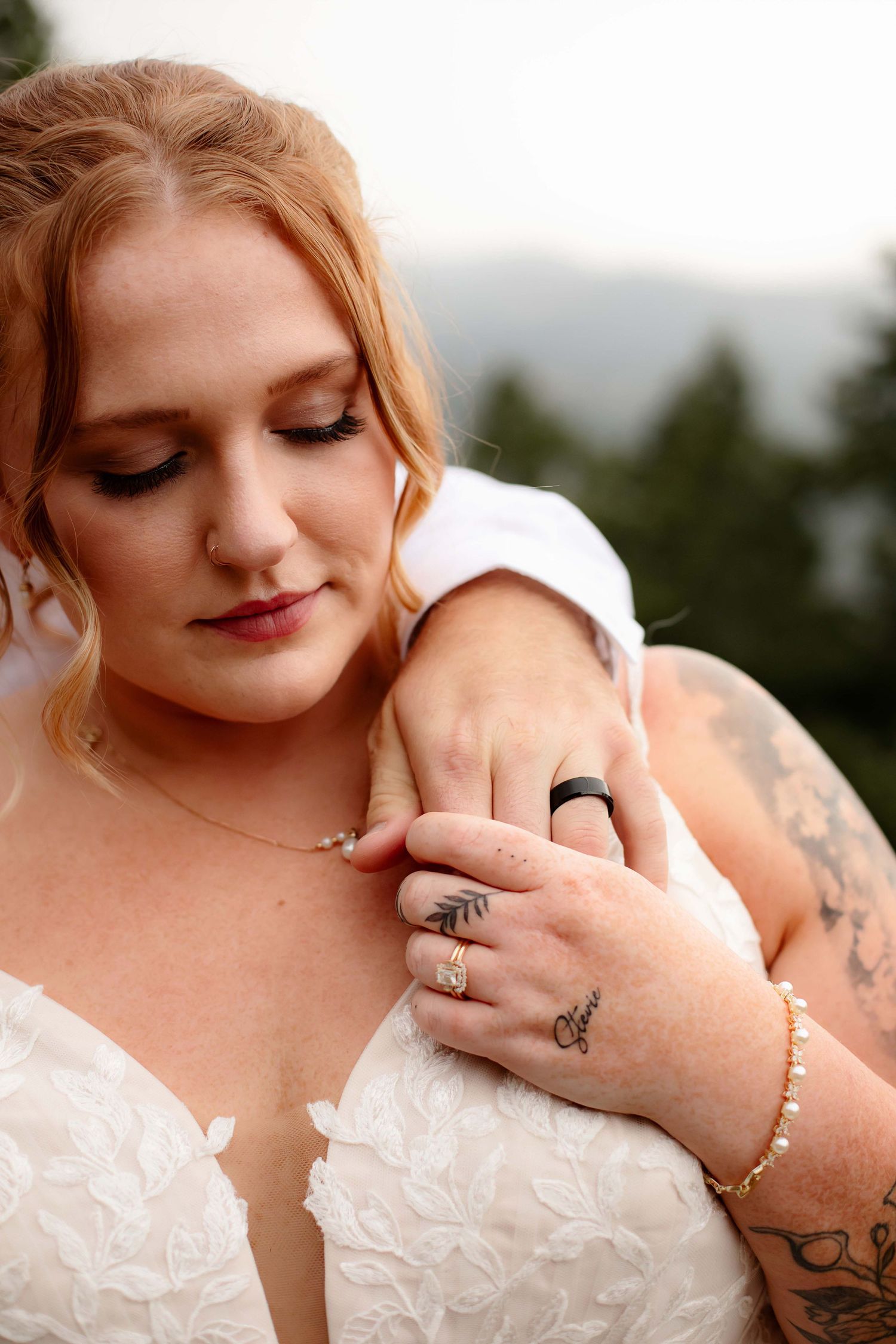 Close-up details of wedding rings and jewelry on hands with delicate lace dress fabric in the background.