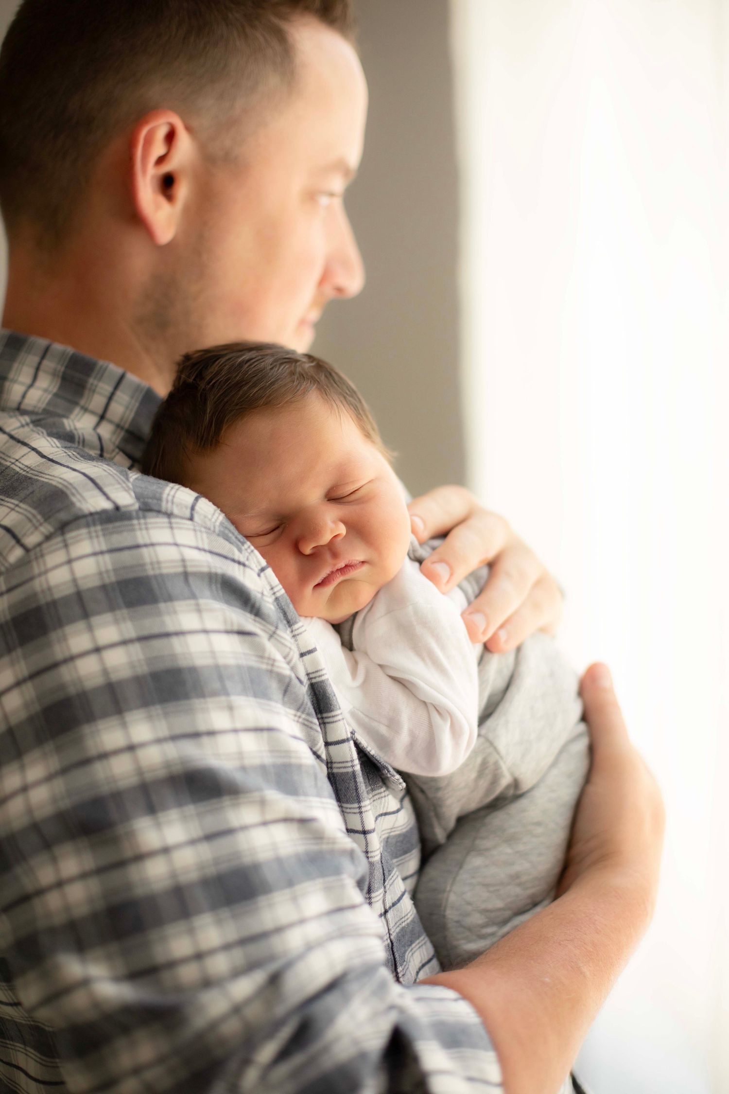 A father gently holds his sleeping newborn baby against his chest, wearing a plaid shirt in a softly lit room.