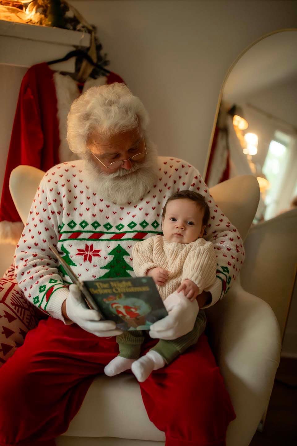 Santa Claus in festive sweater reads a Christmas book to an infant baby sitting on his lap in cozy holiday setting.