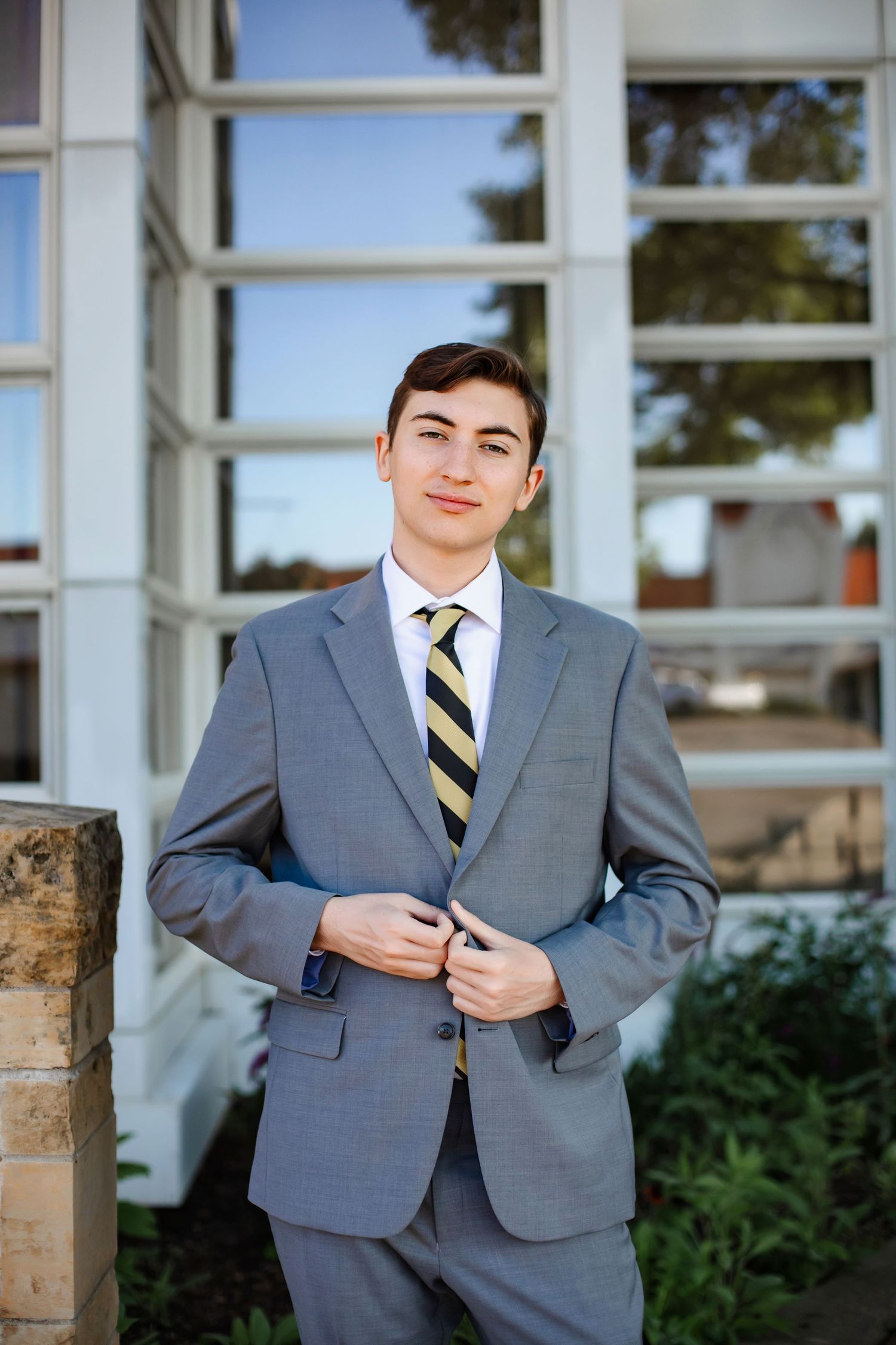 Young man in gray suit and striped tie smiling outdoors near building windows.