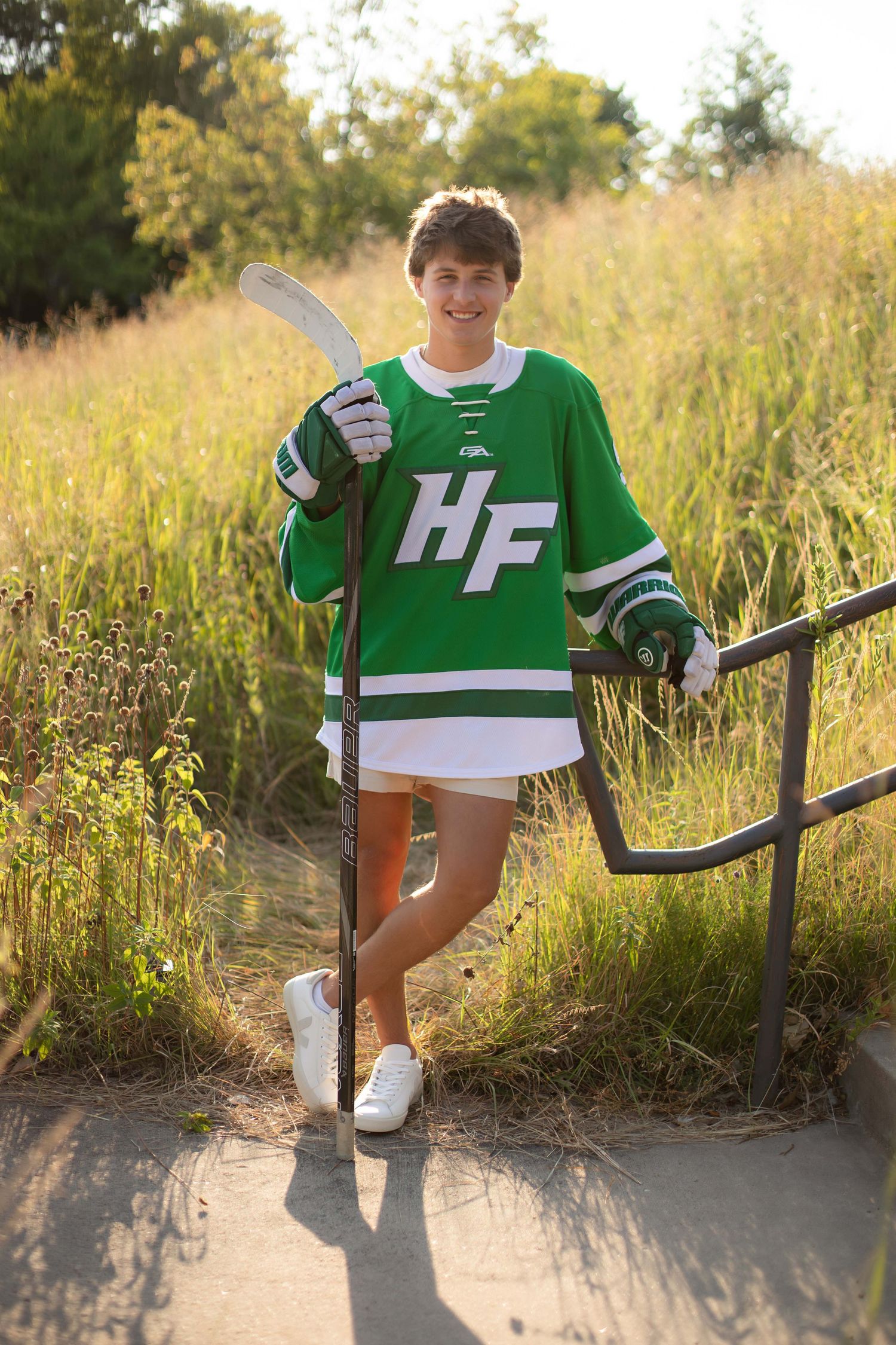 Teen hockey player in green HF jersey holding a stick outdoors in golden hour light.