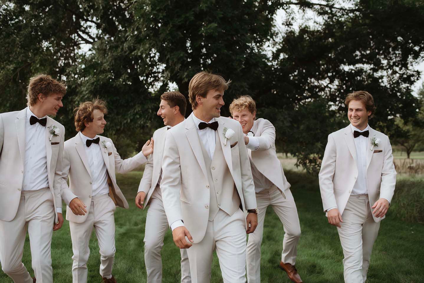 Groom and groomsmen in white tuxedos with black bow ties walking outdoors at a wedding celebration.