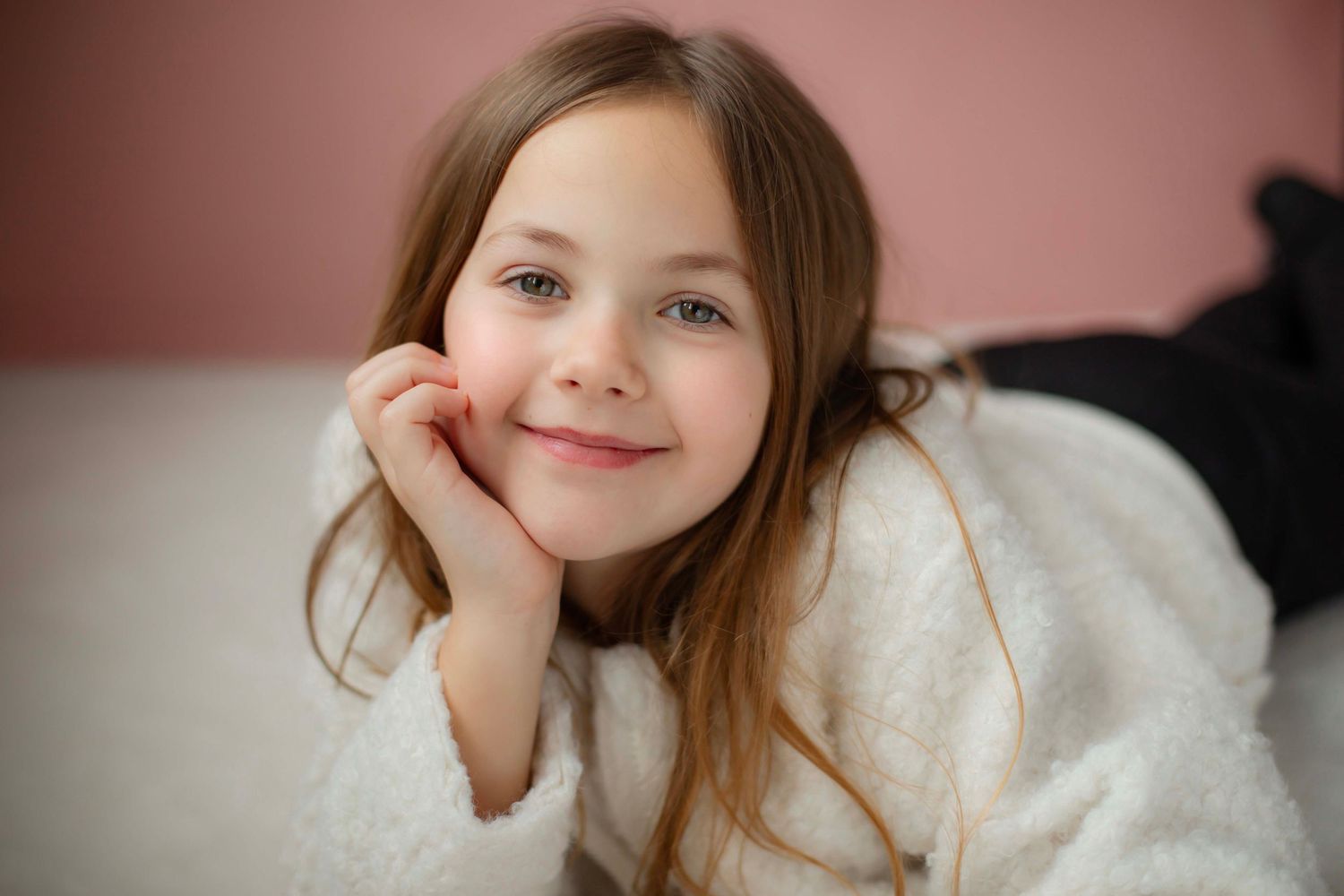 A young child with long brown hair and a sweet smile rests their head on their hand while wearing a white sweater.