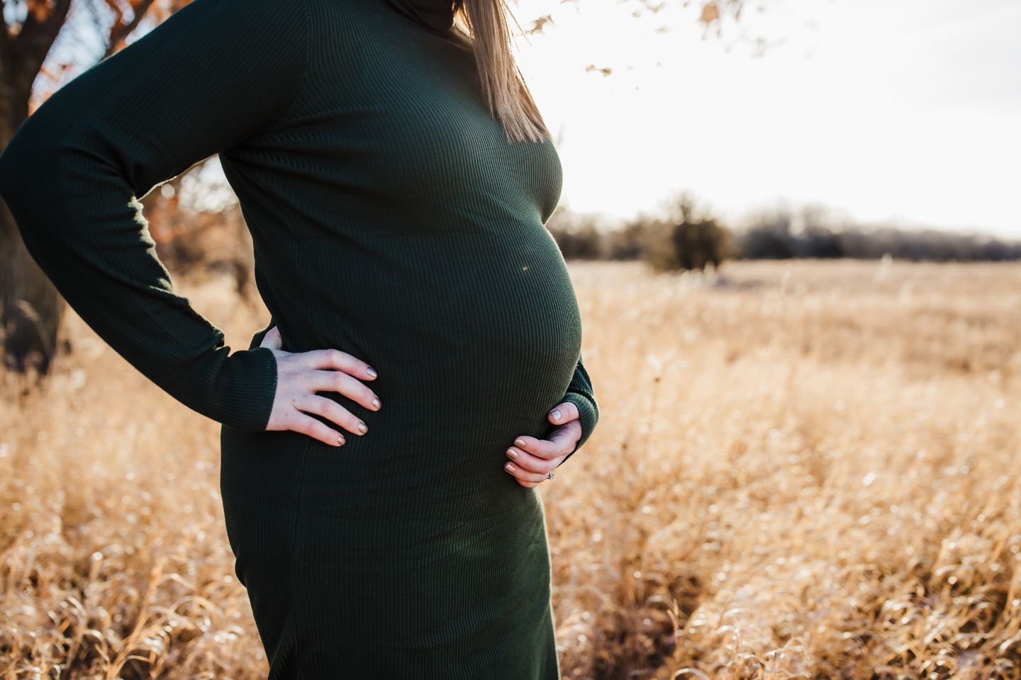 Silhouette of pregnant woman in black dress holding her belly in golden wheat field at sunset.