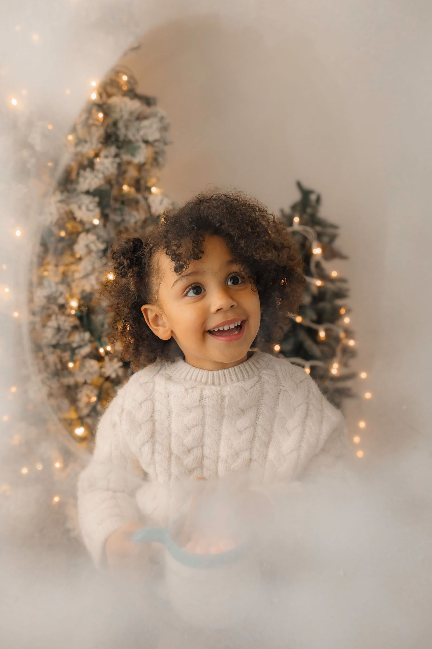 Cozy holiday portrait with child in white sweater smiling by illuminated Christmas trees.