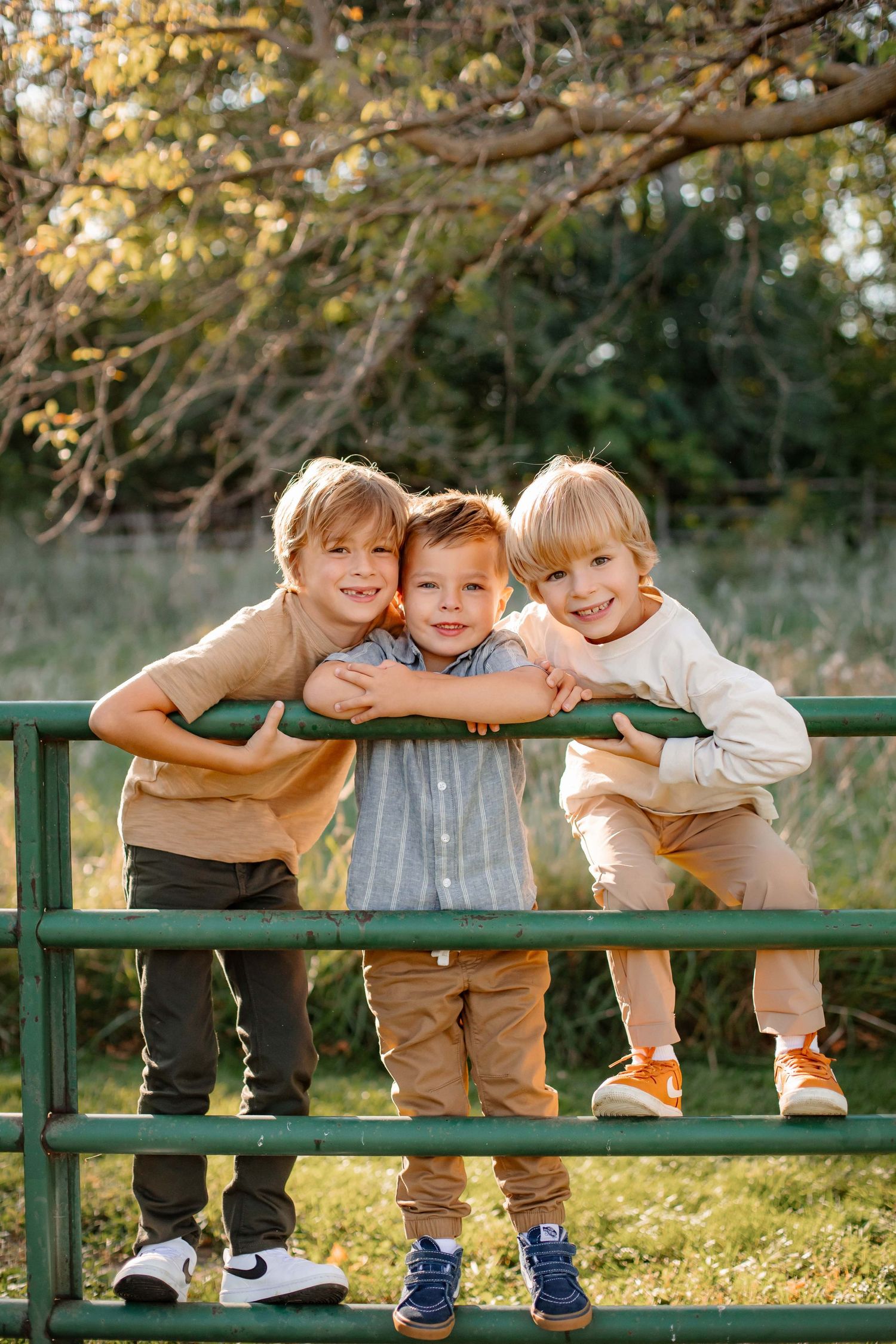 Three young children lean on green railing outdoors with bright smiles.