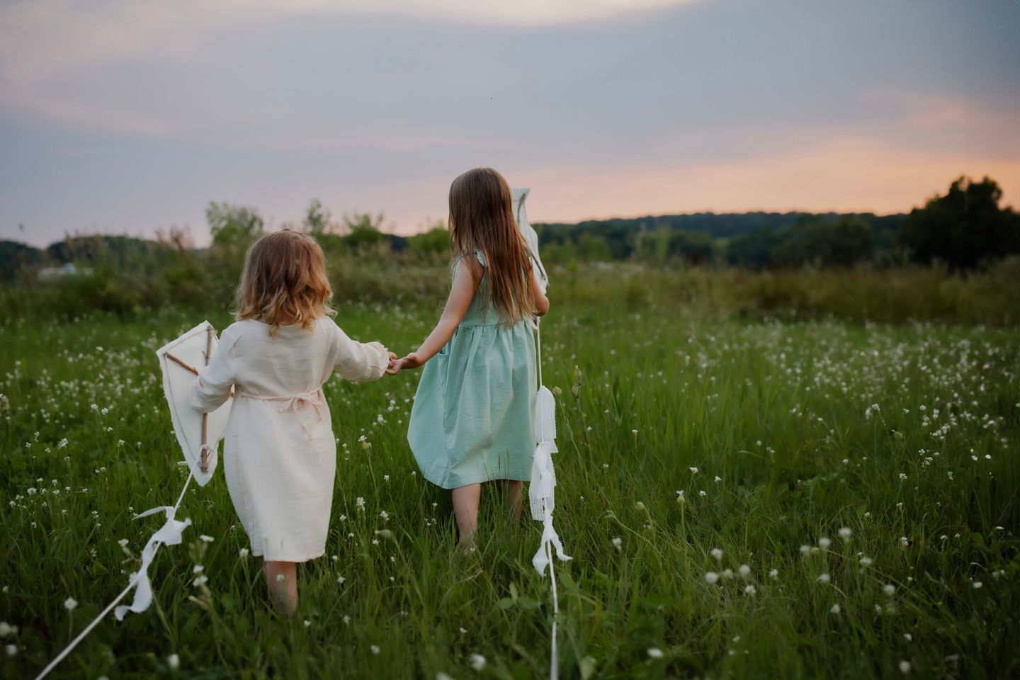 Two children holding hands while running with kites through a flowering meadow at sunset.