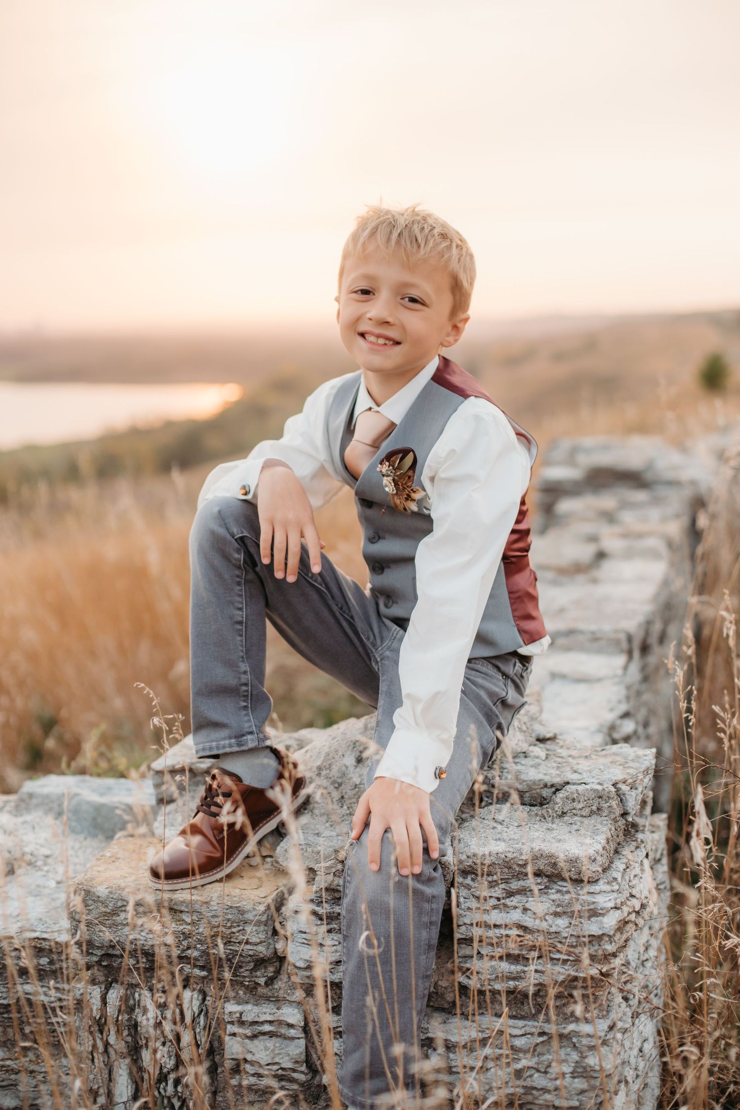 Young boy in formal vest sits on stone wall during sunset photoshoot.