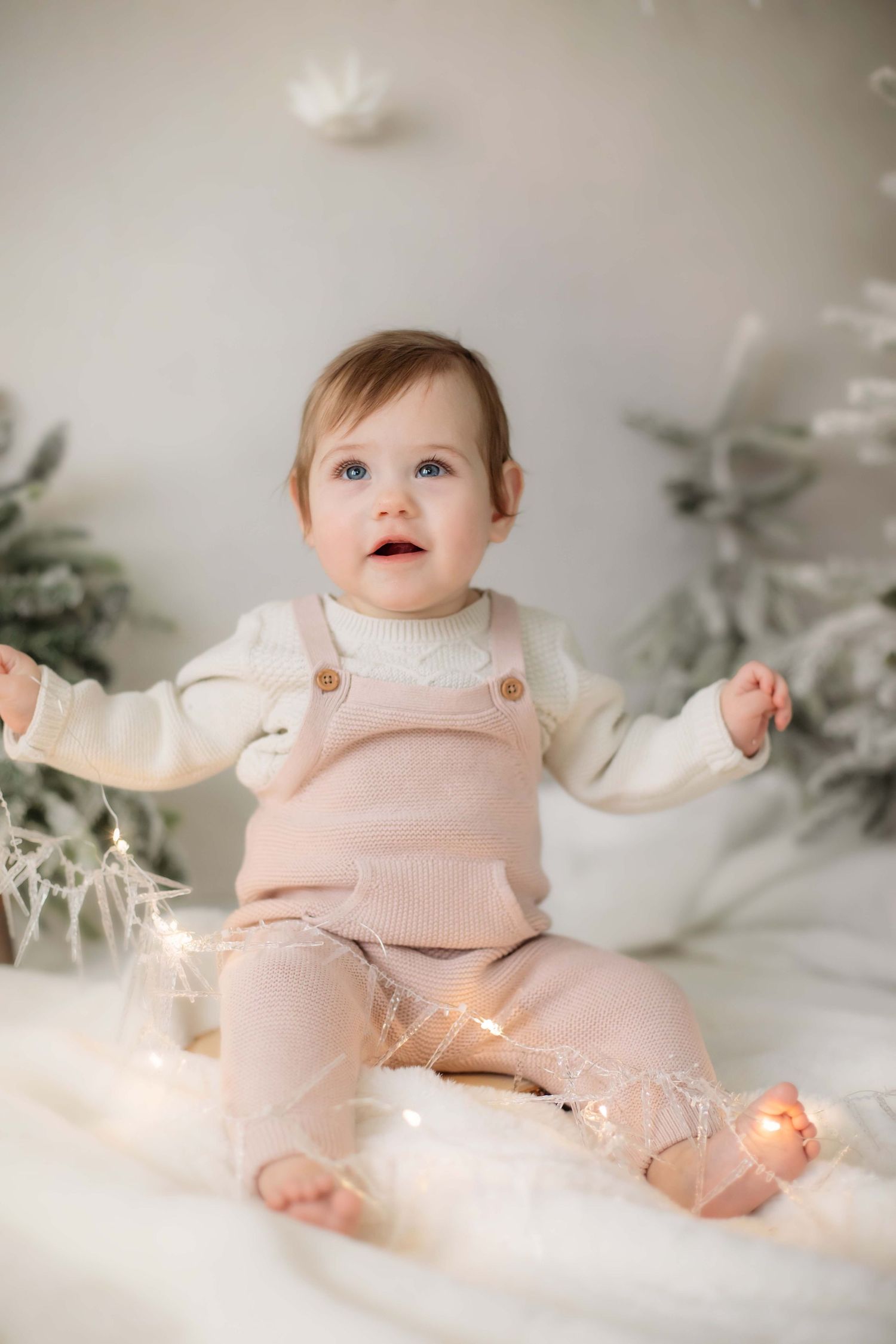 Baby in pale pink overalls and white sweater sits among twinkling lights and winter decorations.