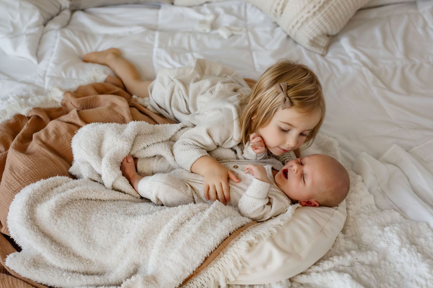 Cozy sibling cuddles in cream-colored knit sweaters on white bedding.