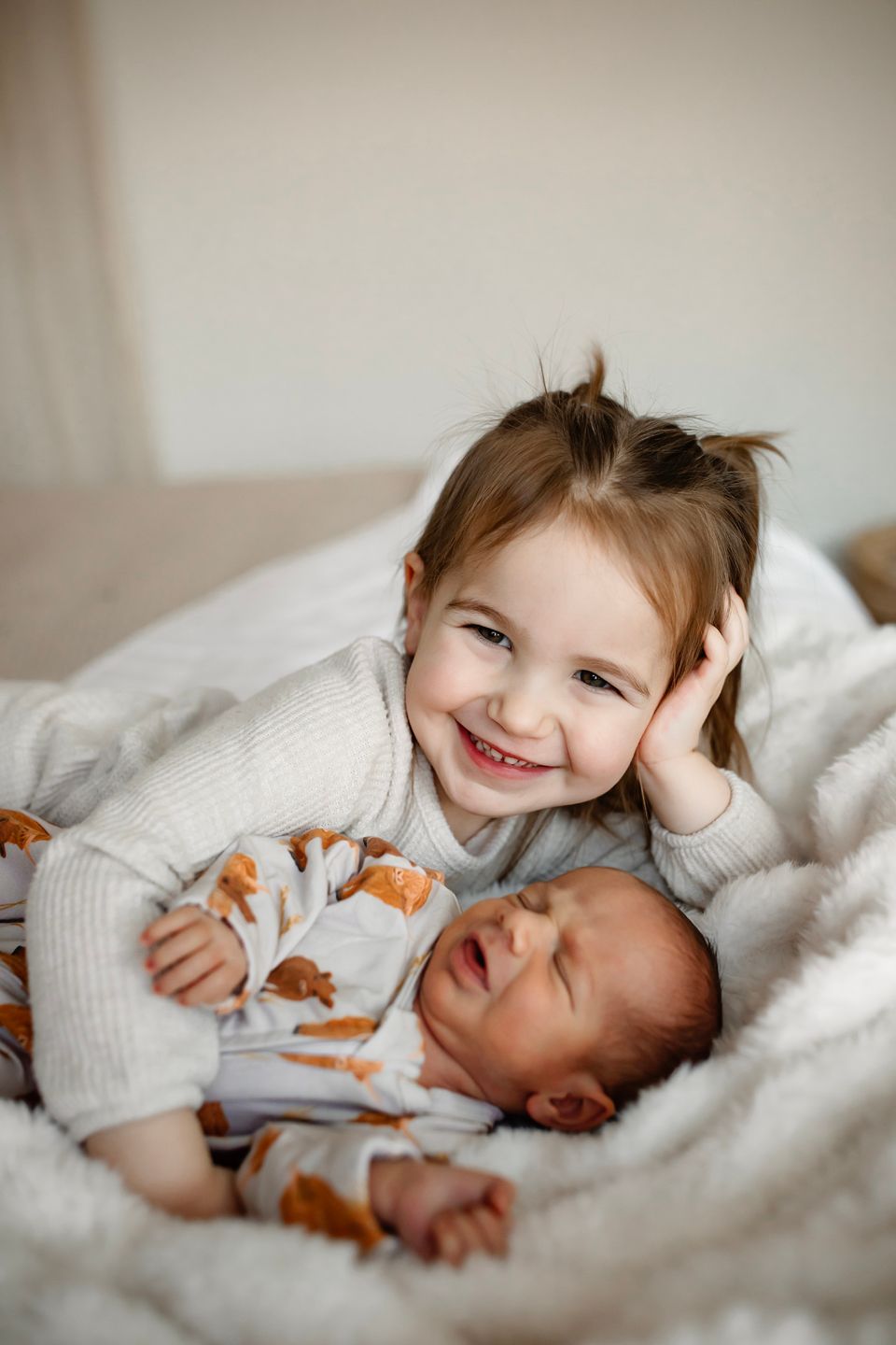 Two siblings share a tender moment lying together on white bedding, with the older one smiling joyfully.