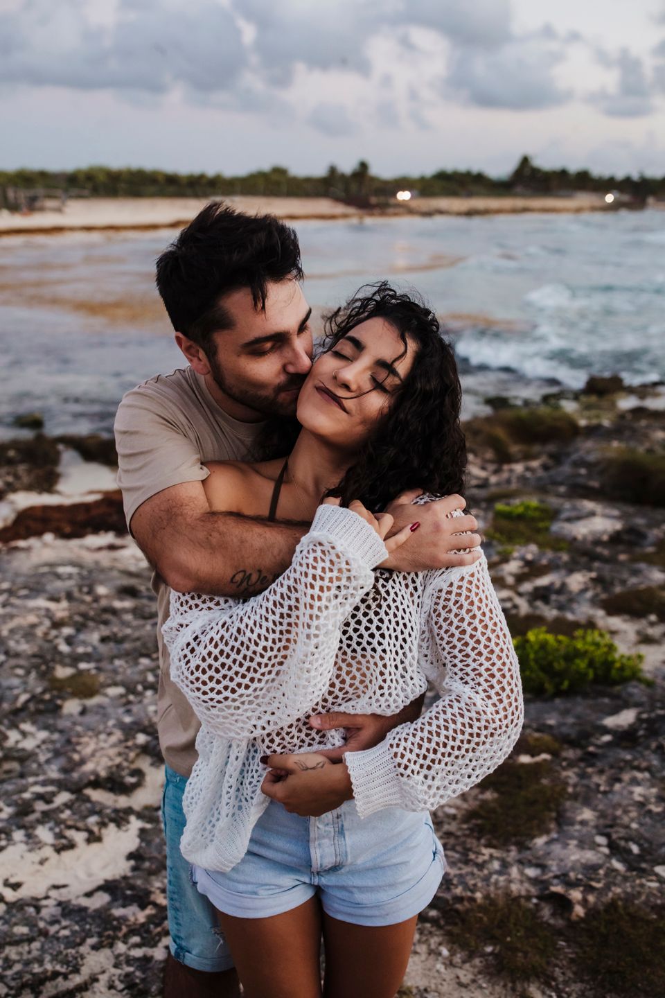 Romantic couple embrace on rocky shoreline during golden hour.