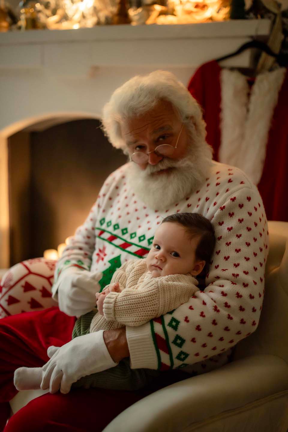 Santa Claus holding a baby infant on his lap in front of a cozy fireplace during Christmas.