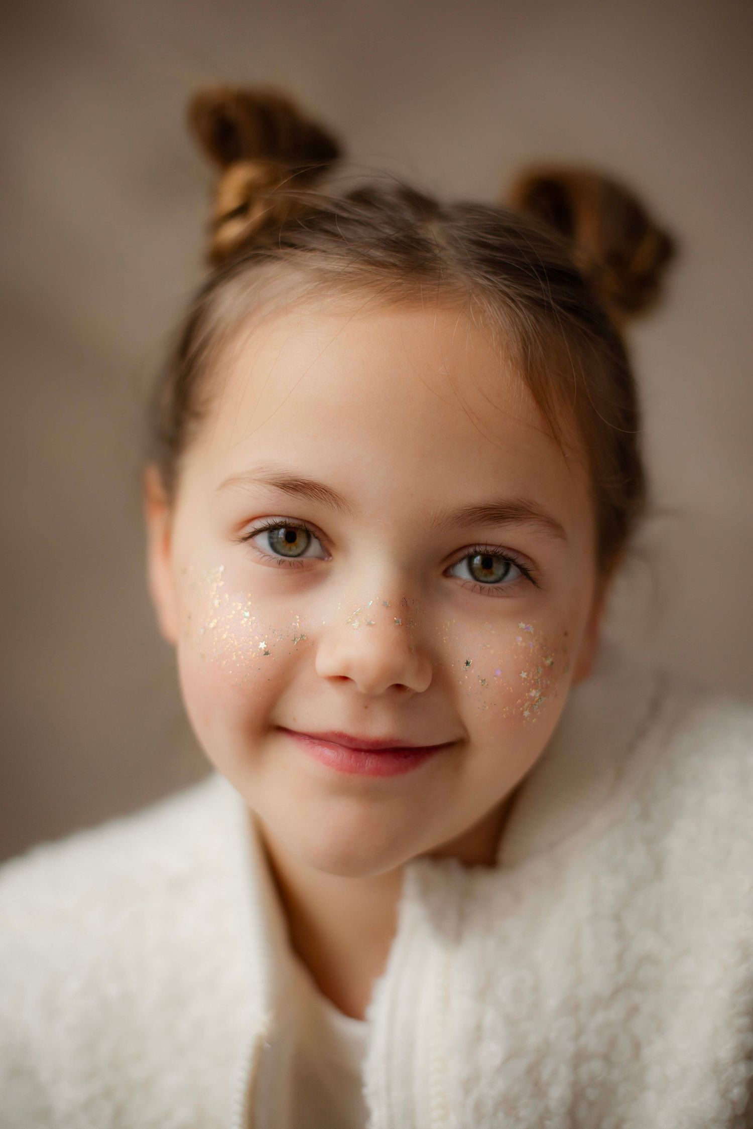 Studio portrait with sparkly face decorations and a cozy white sweater against a neutral backdrop.