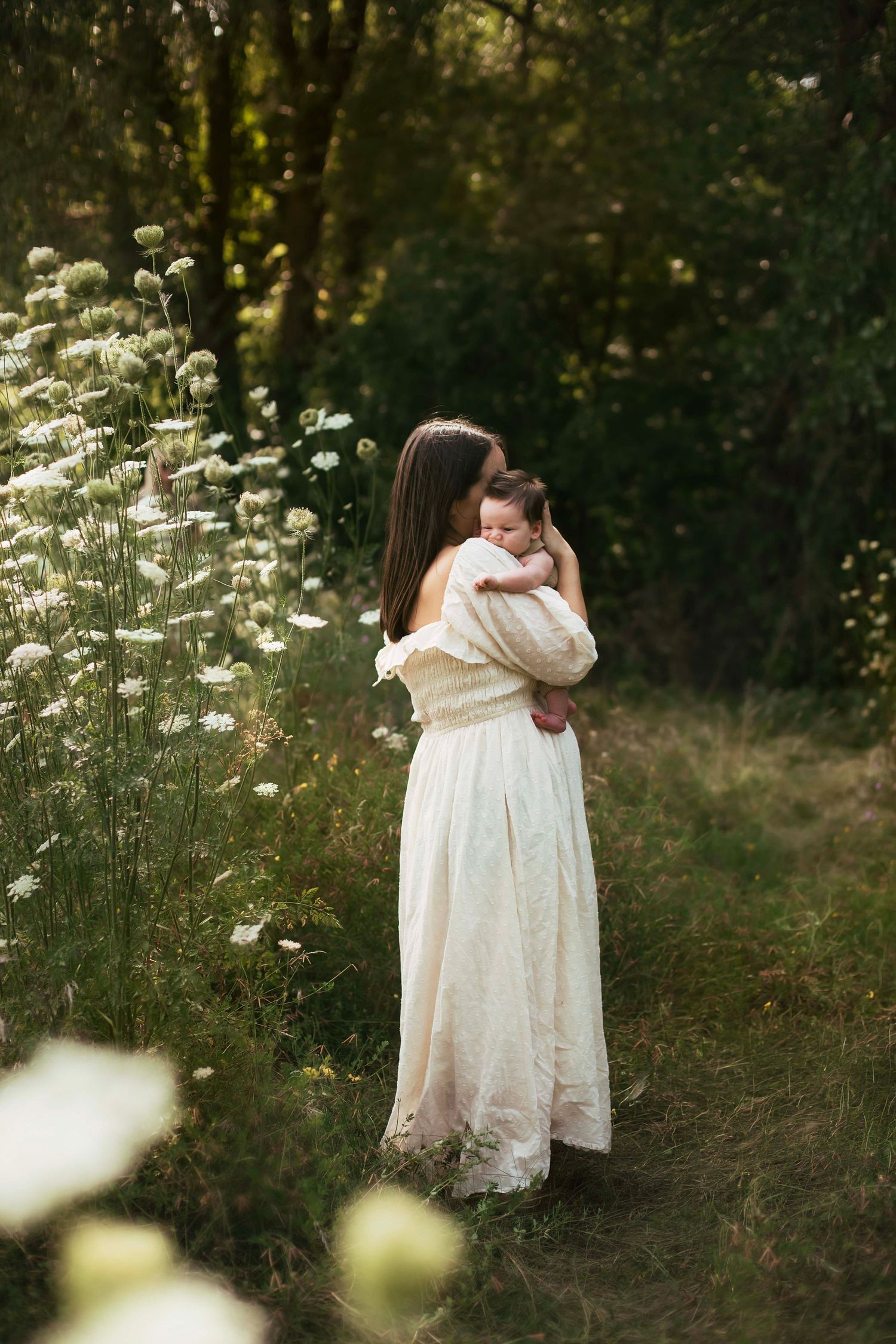 A woman in a white dress holds a baby among wildflowers in a lush green forest setting.