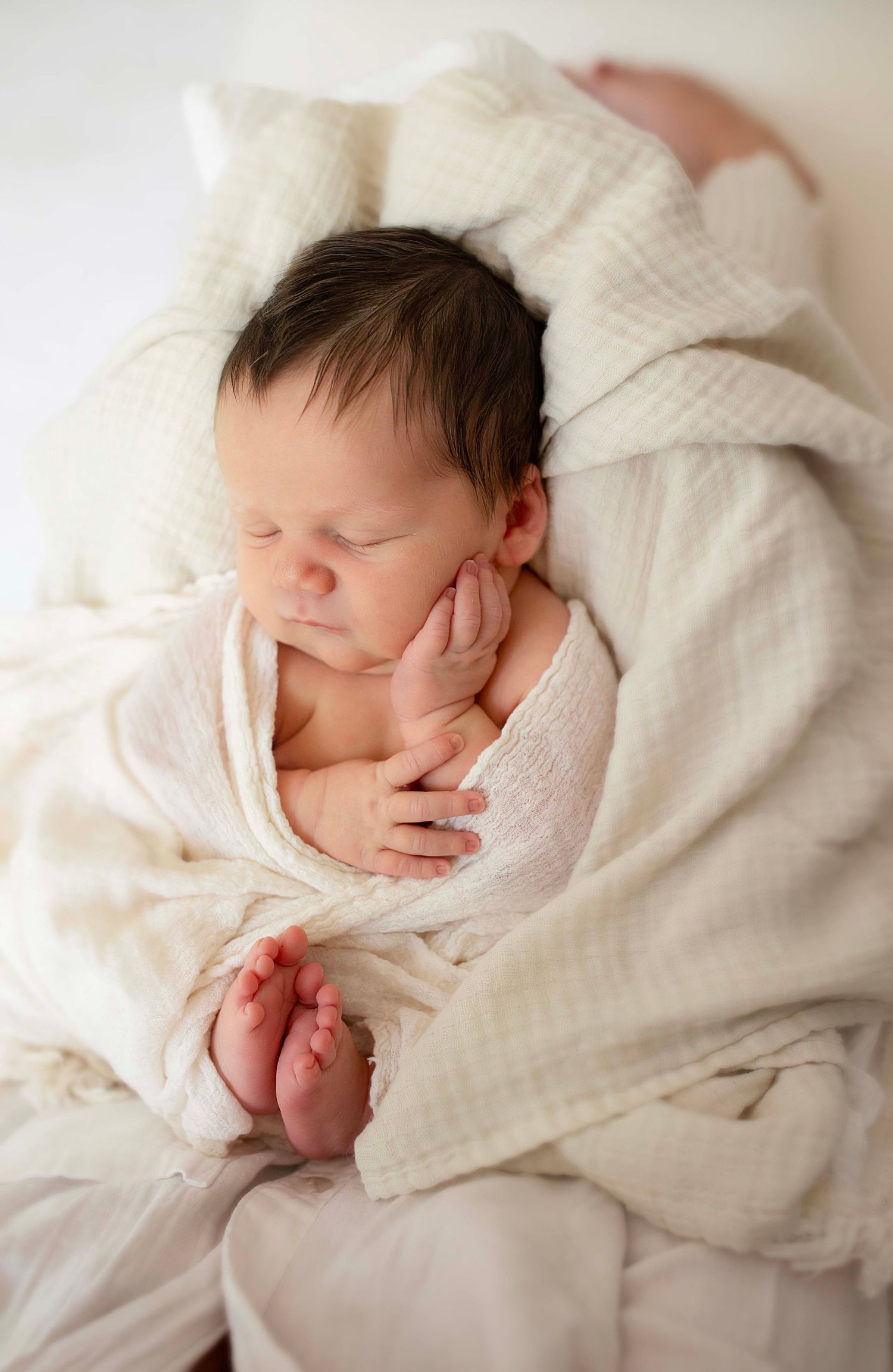 Peaceful newborn baby sleeping wrapped in a soft white muslin blanket with tiny toes peeking out.