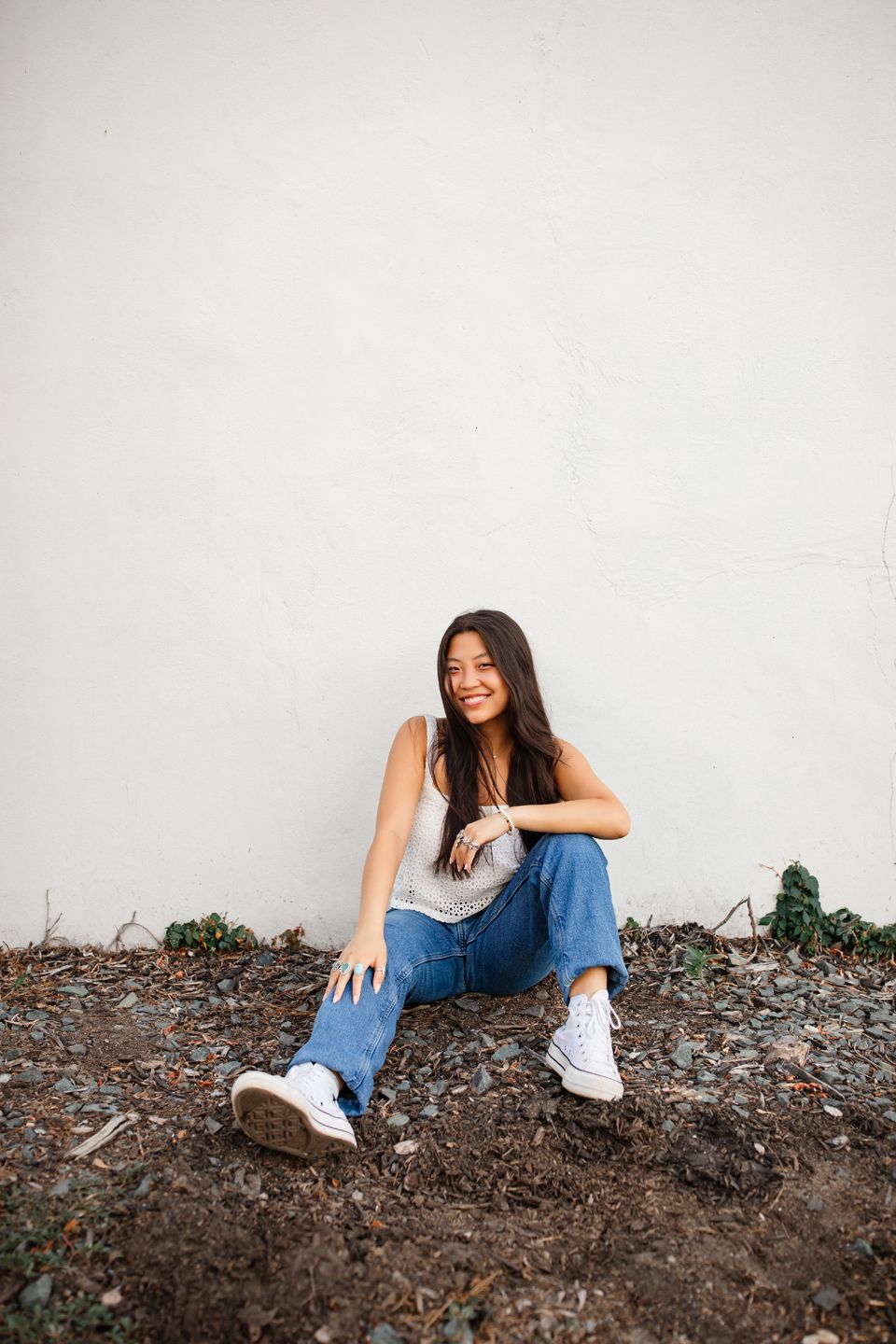 Casual portrait of someone sitting on ground wearing blue jeans, white top and sneakers against white wall.