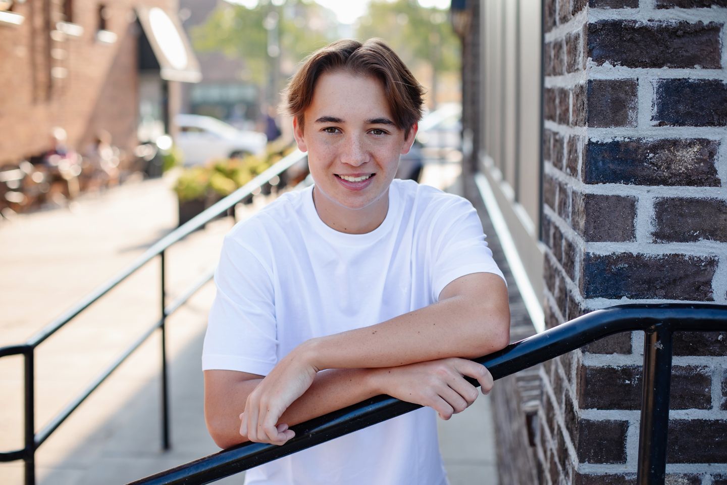 A young person in a white t-shirt leans against a railing by a brick building on a sunny day.