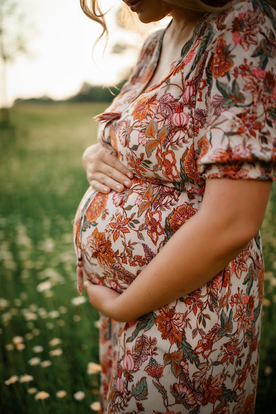 A pregnant figure in a floral dress tenderly holds their baby bump while standing in a sunlit meadow.