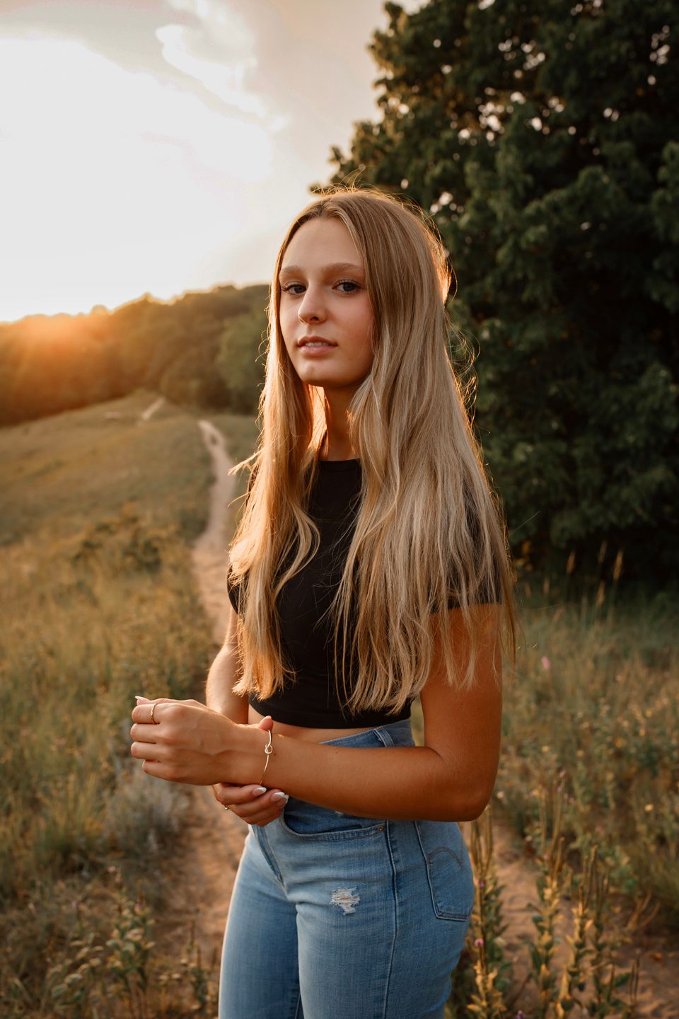 Person in jeans and dark top standing in golden hour light with rolling hills in background.