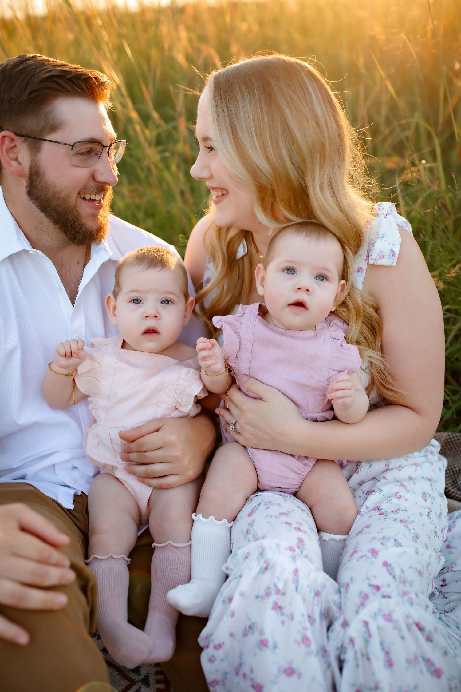 Intimate family portrait with twin babies in pink outfits against sunset-lit field.