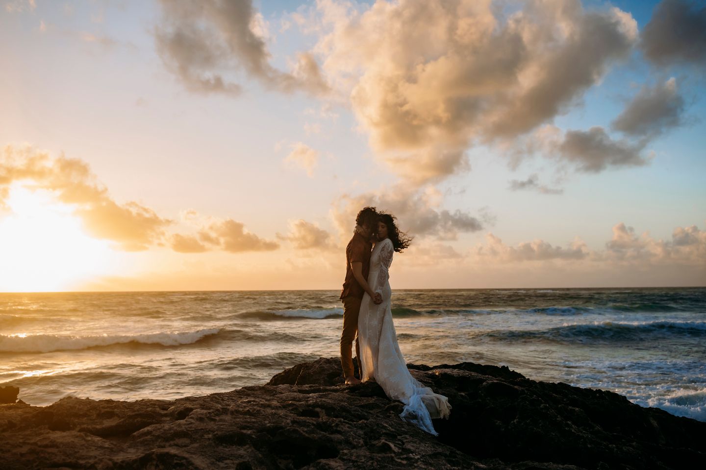 A couple embraces on rocky cliff edge during golden hour with dramatic ocean waves below.