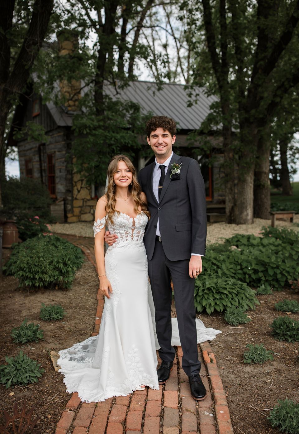 Wedding couple standing on brick path in front of rustic cabin surrounded by trees.