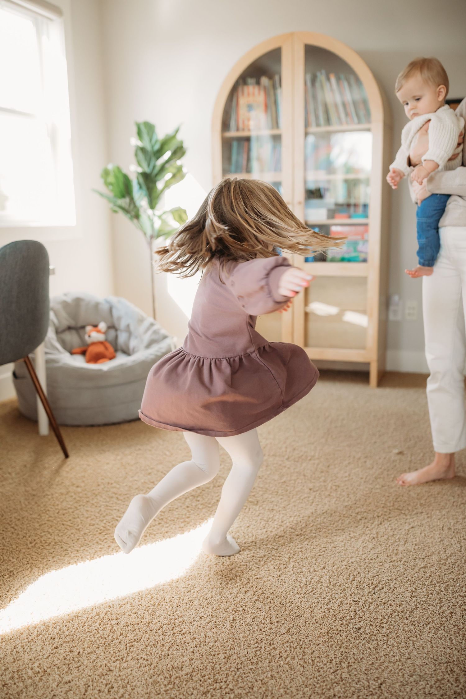 A child in a pink dress twirls in a sunlit room while playing.