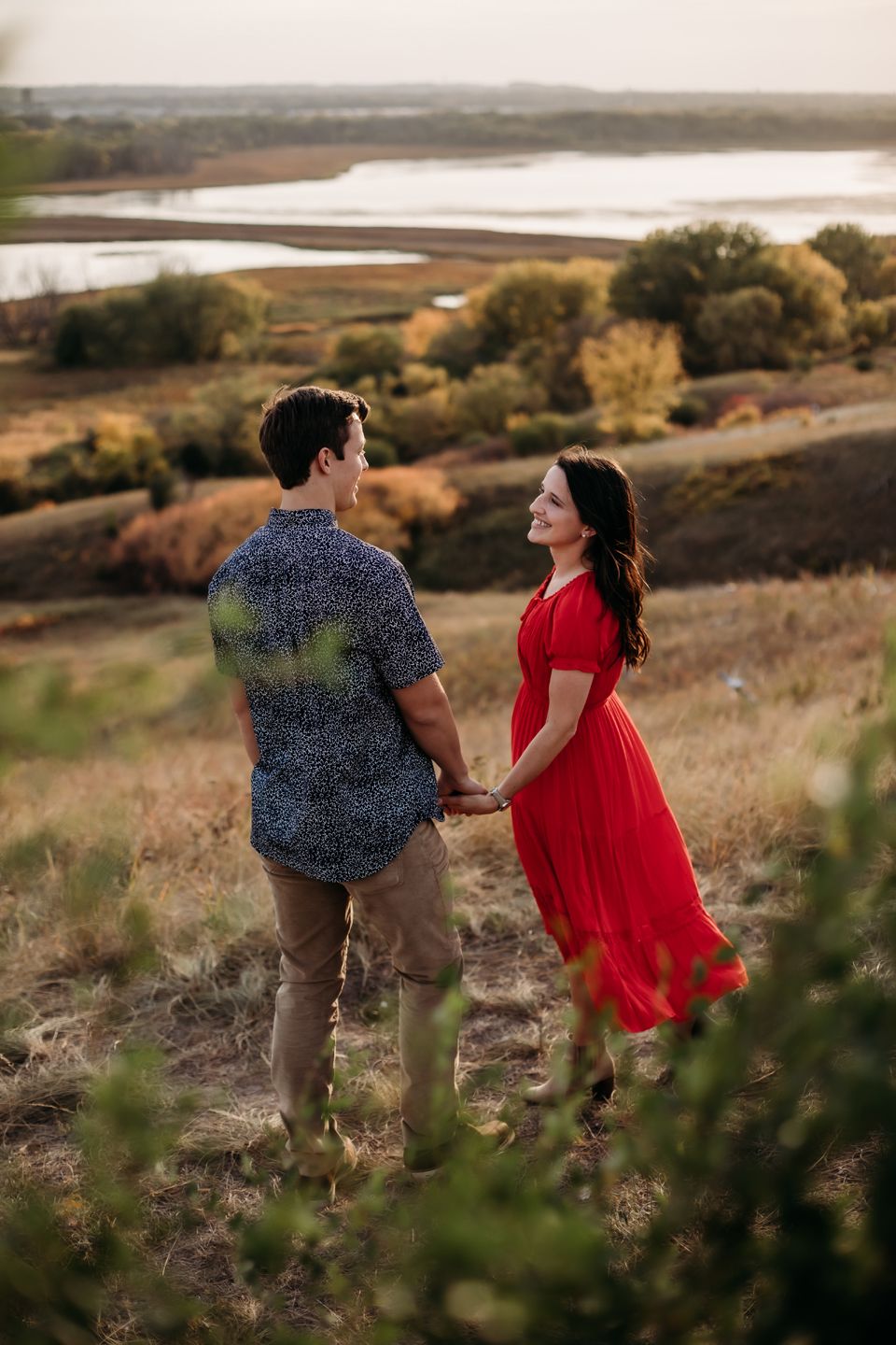 A romantic couple holds hands on a hilltop overlooking a scenic lake during golden hour.