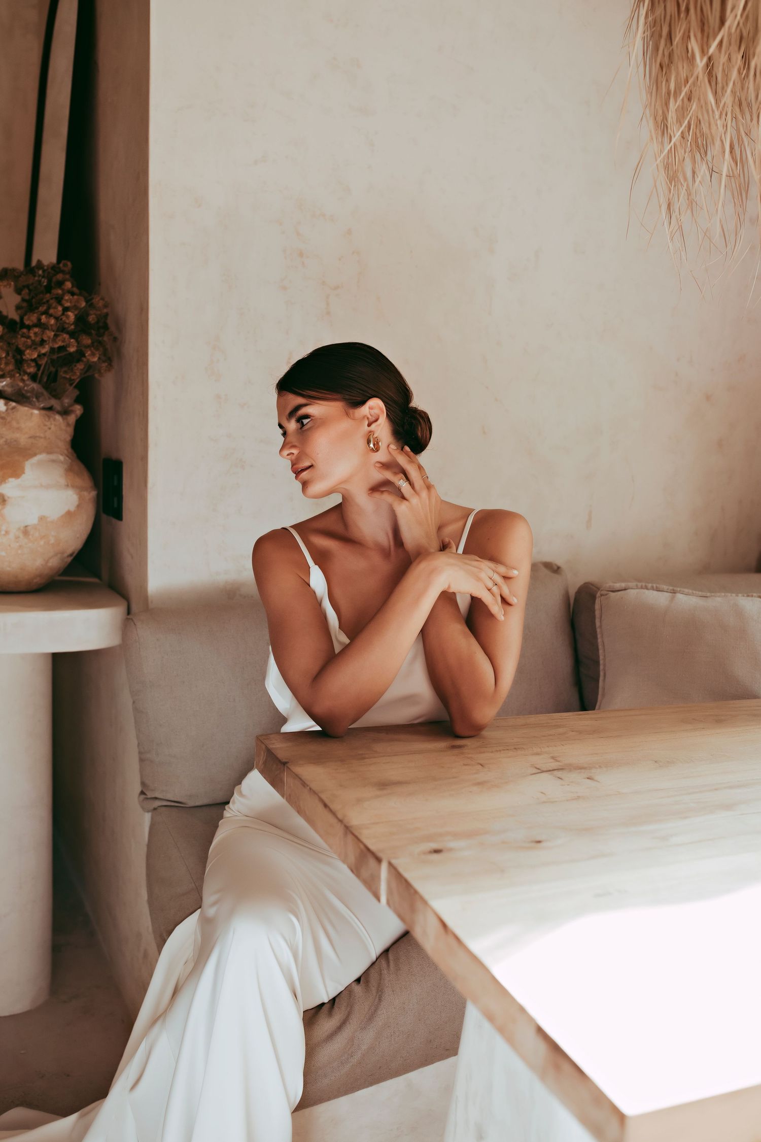 Woman in white wedding dress poses elegantly at wooden table in minimalist interior with soft natural lighting.