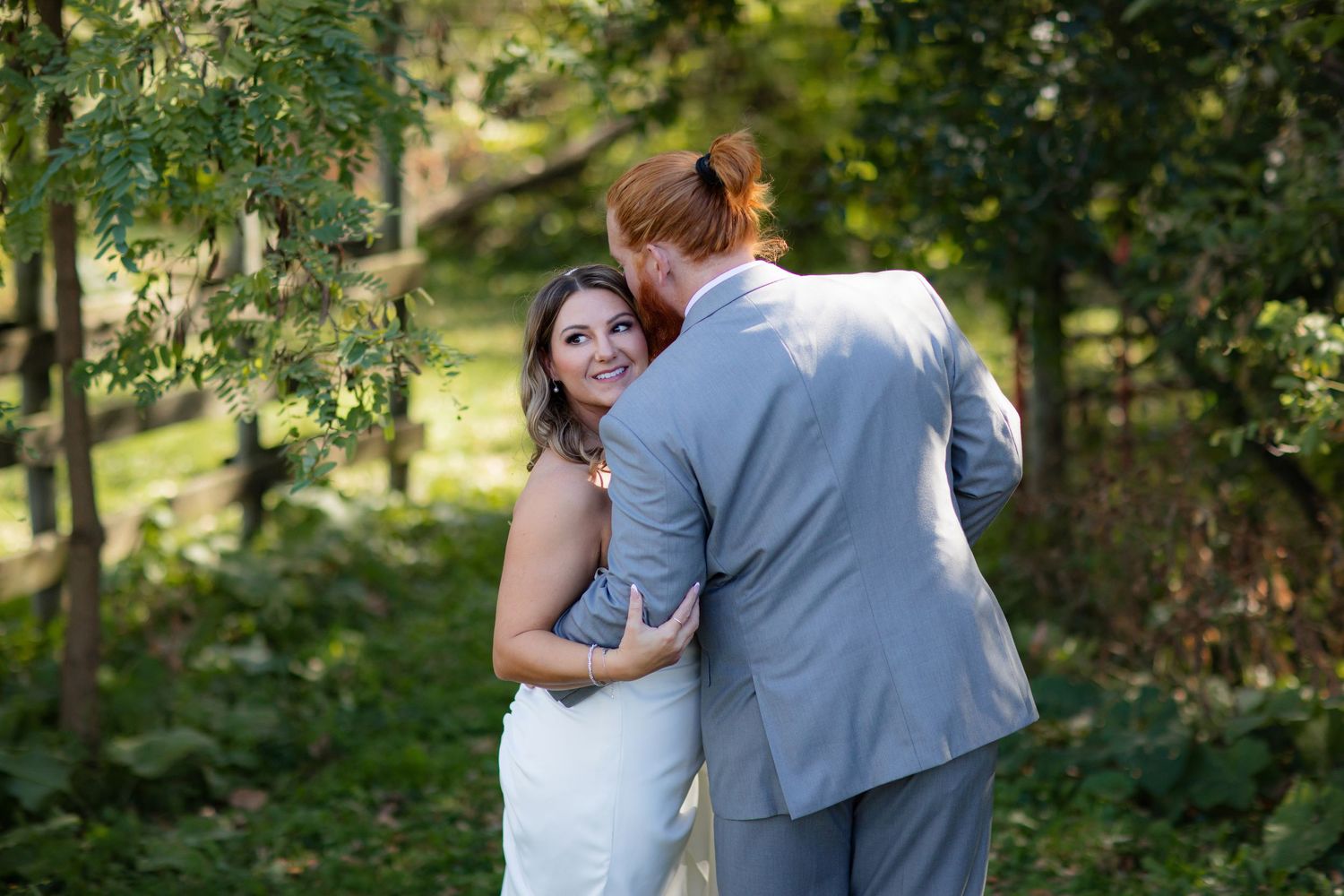Intimate wedding couple embracing in a lush garden setting with sunlight filtering through trees.