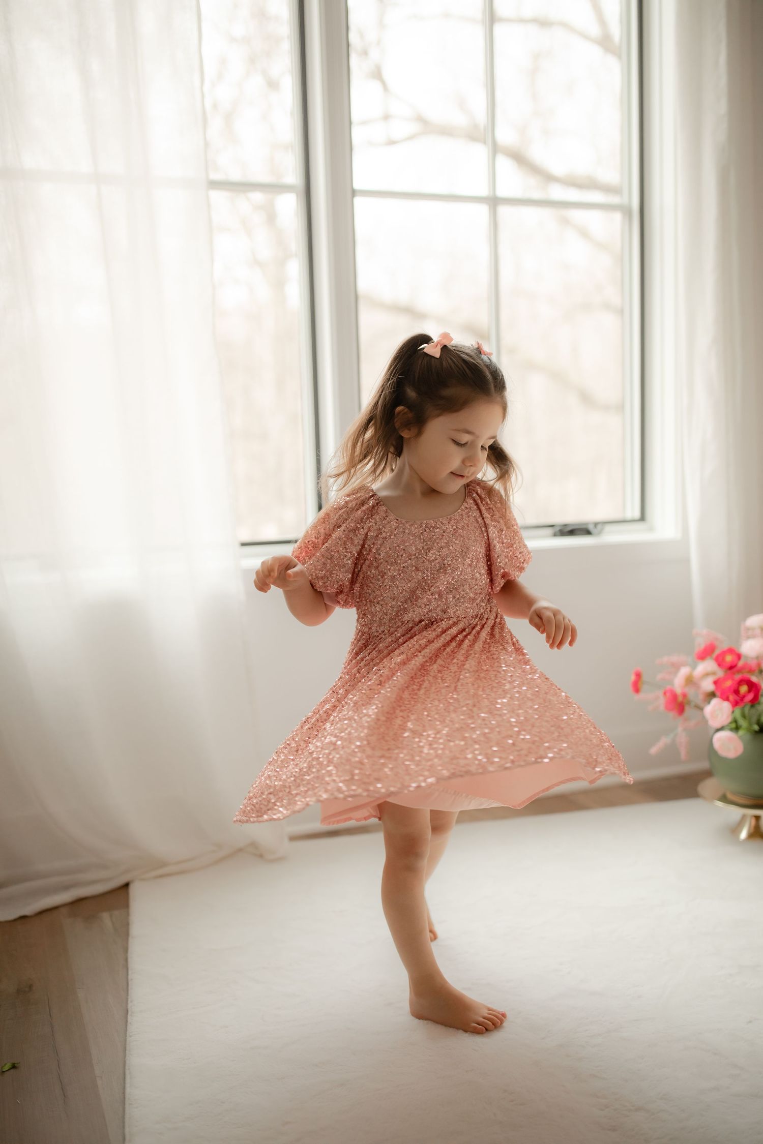 A young girl in a pink lace dress twirls barefoot by a sunlit window with flowers nearby. Willow & Wren Photography studio in Chaska, MN.
