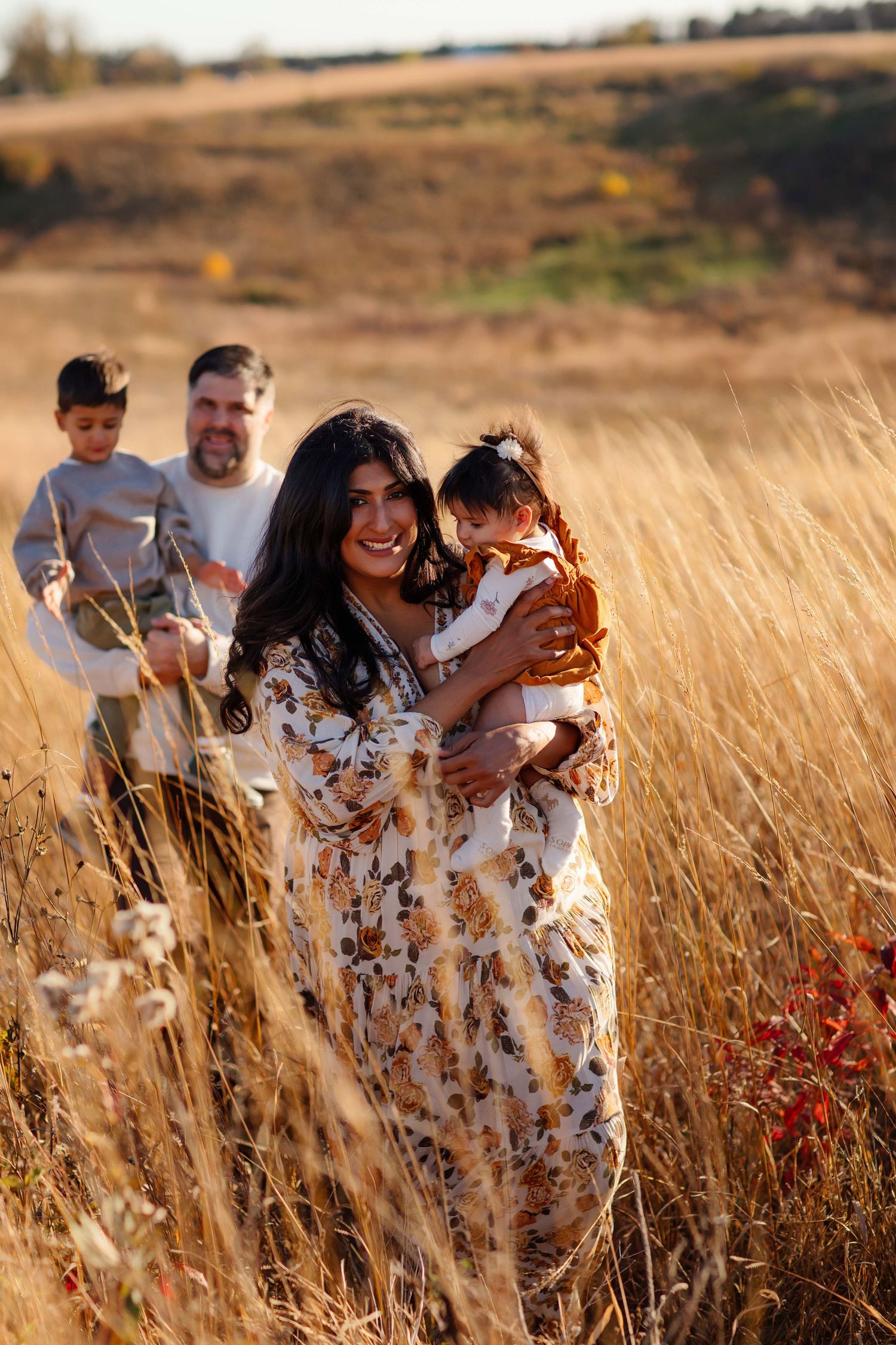 Family walking in golden autumn field, mother holding baby in rust outfit, father carrying son in background.