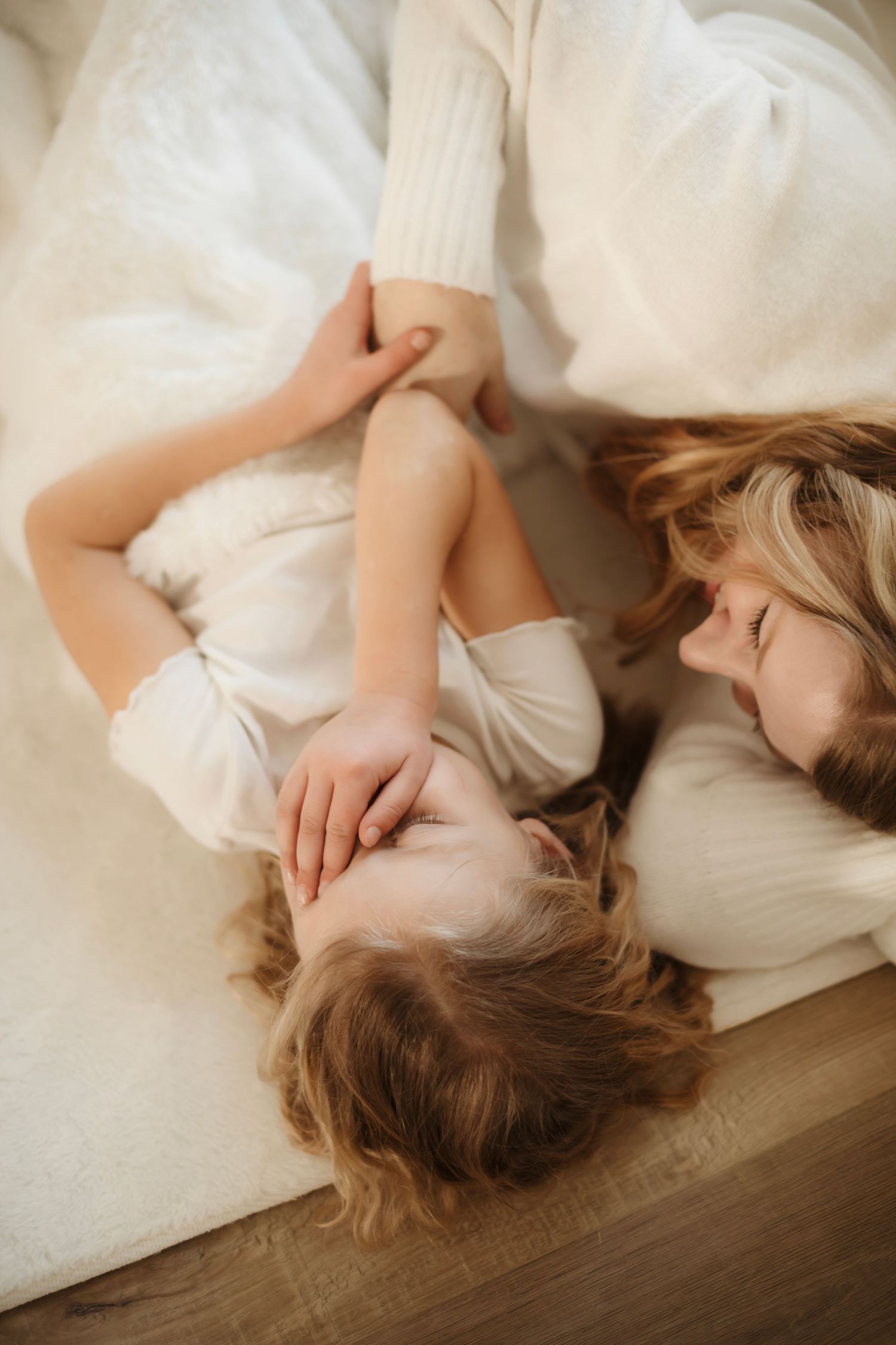 Person lying in white bedding with hair spread on wooden floor in artistic boudoir photography series.