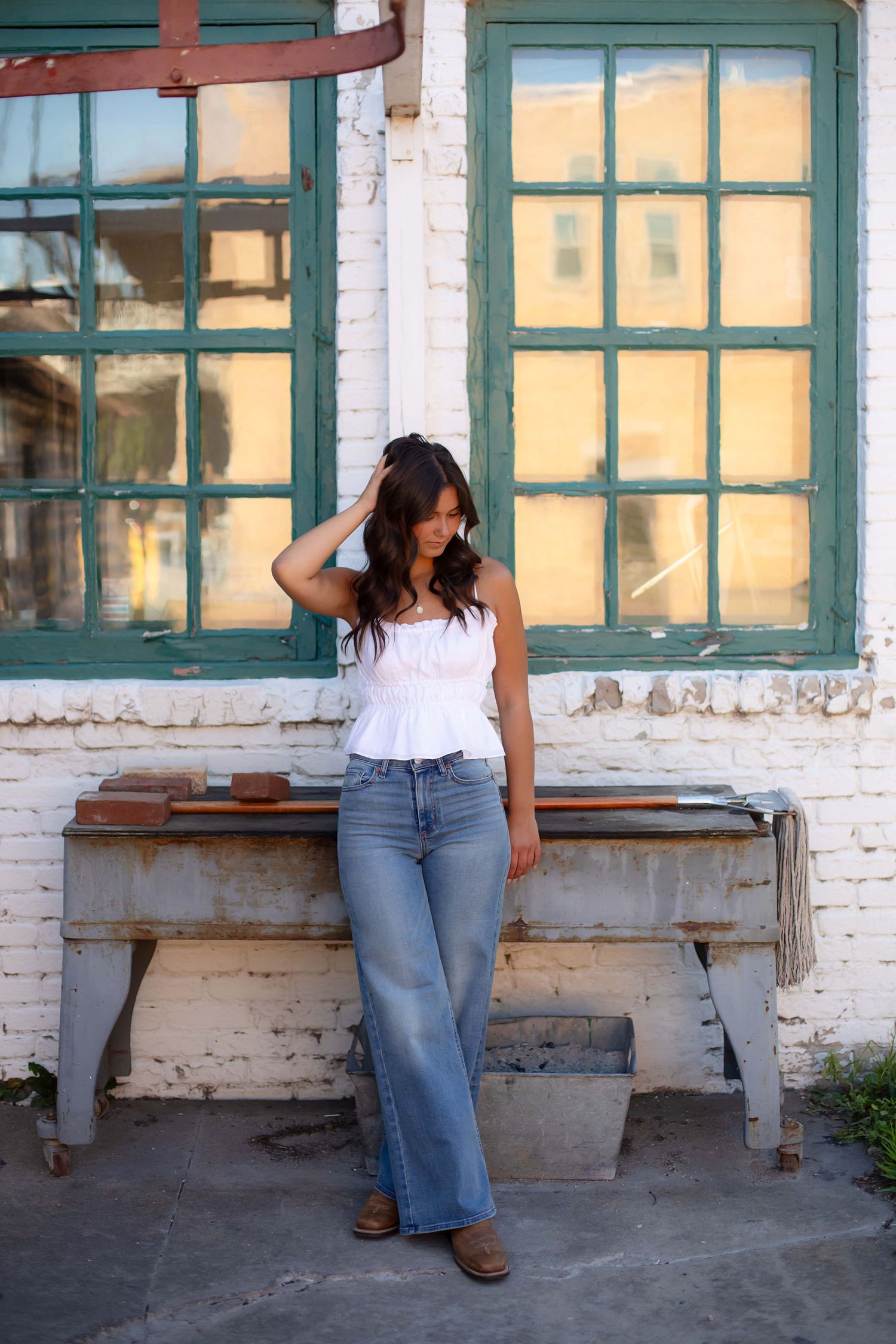 Woman in white peplum top and wide-leg jeans leaning against vintage workbench outside whitewashed brick building.