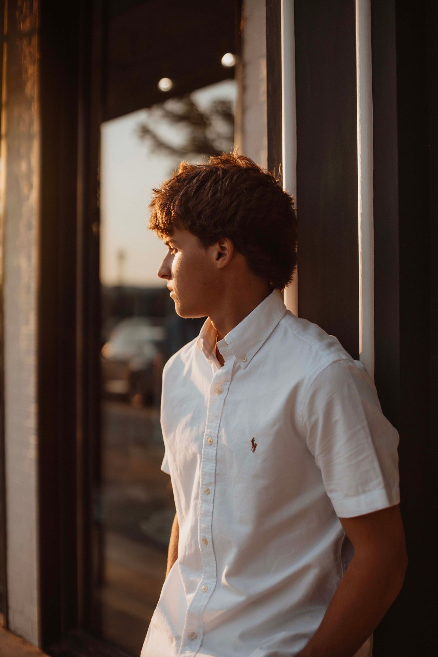 Teen boy in white Ralph Lauren shirt leaning against door frame in golden light.