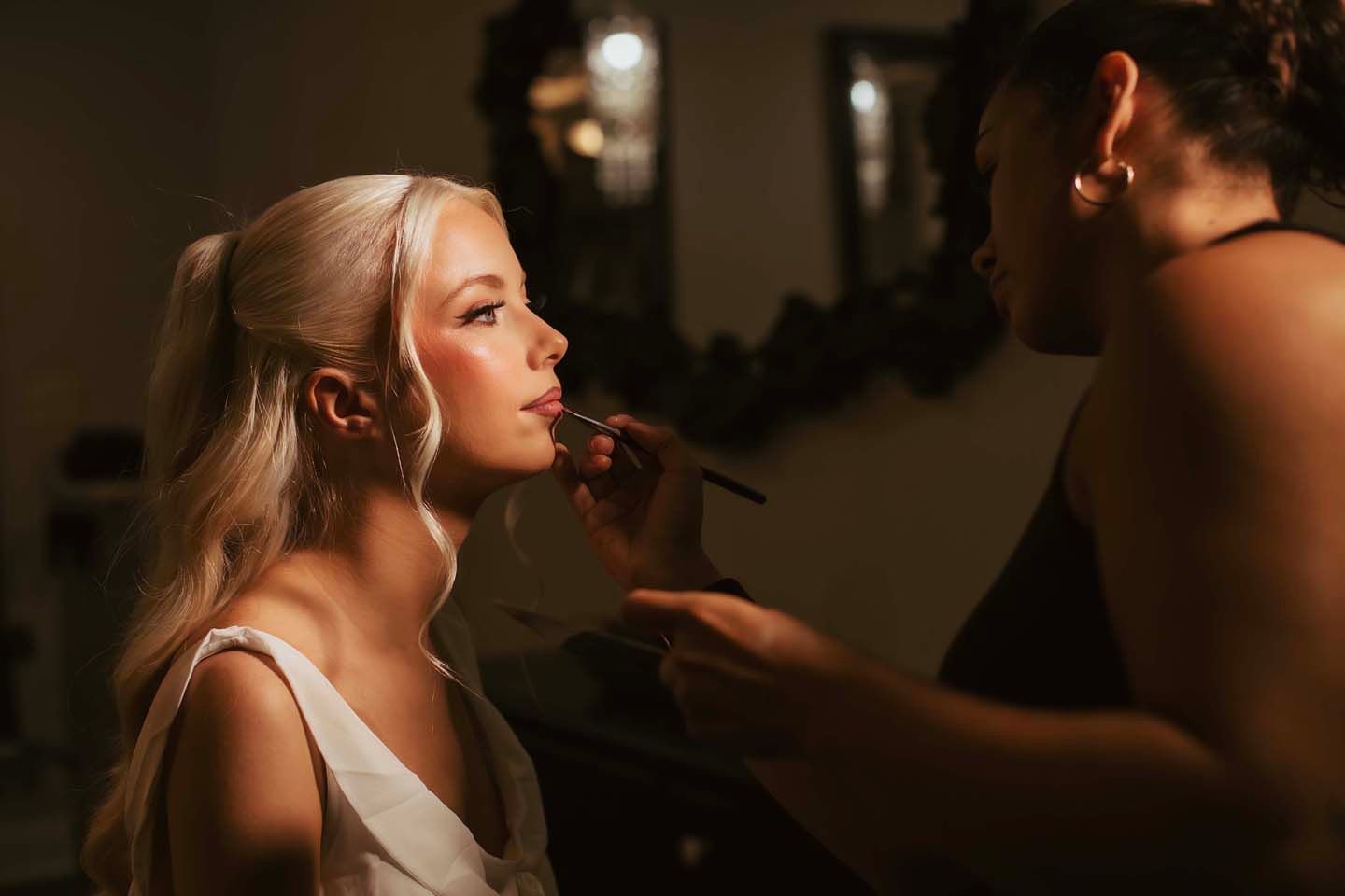 A makeup artist applying blush to a blonde woman's face with a brush in a warmly lit studio.