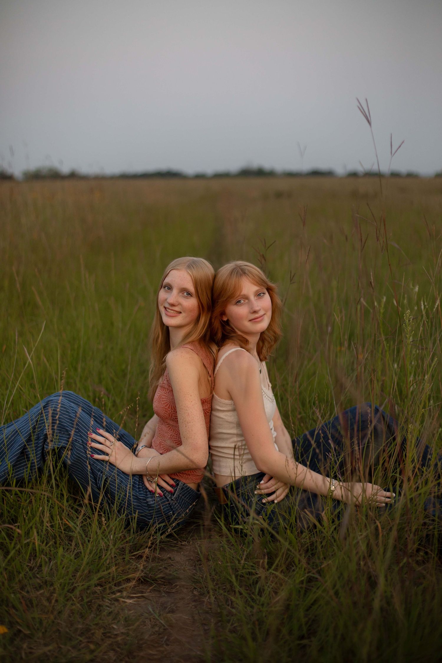 Two redhead girls sitting back-to-back in a green field, wearing jeans and tank tops, smiling outdoors at dusk.