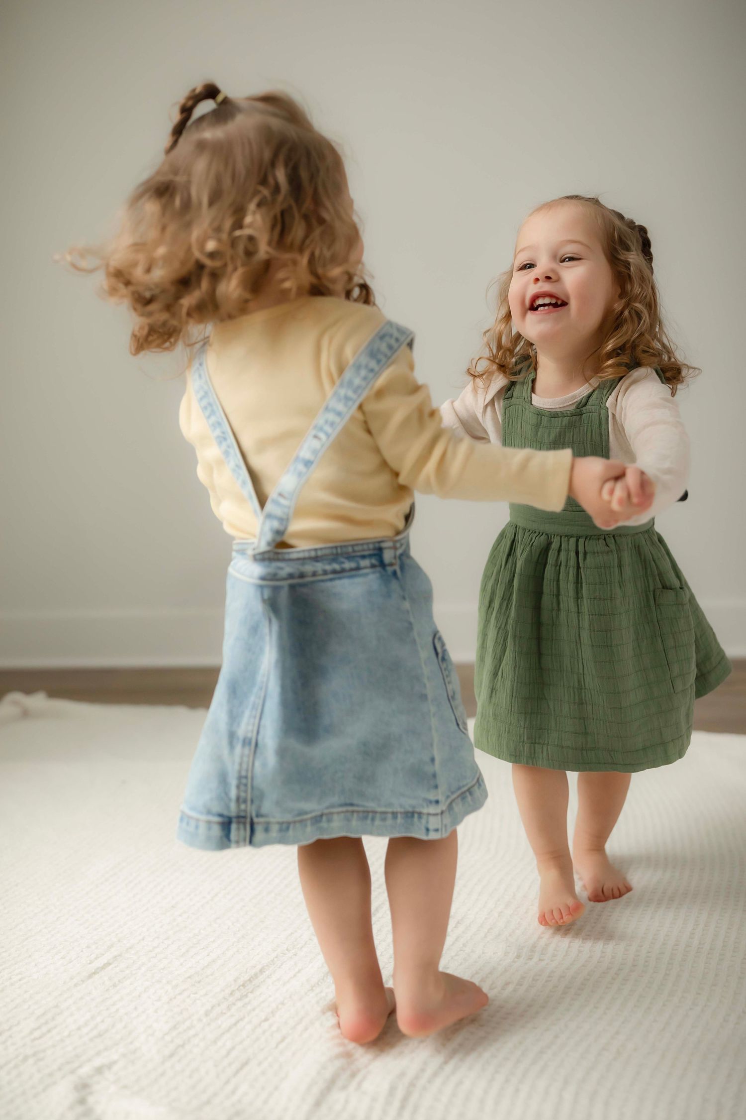 Two toddlers in denim and green dresses dance and play together on a white carpet.