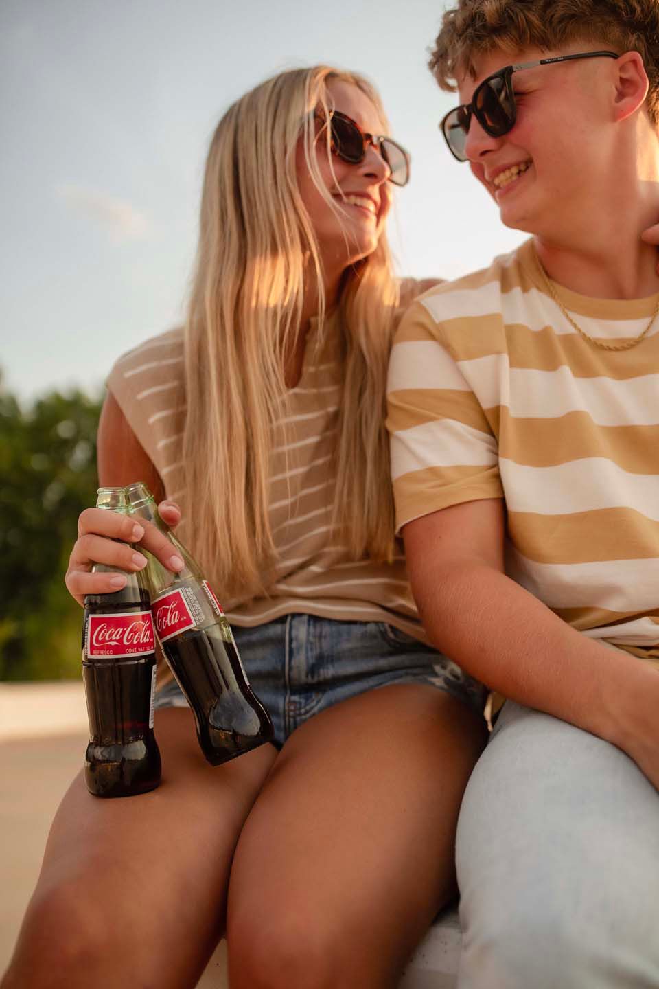 Two smiling teens in striped shirts share Coca-Cola bottles while sitting together outdoors at sunset.